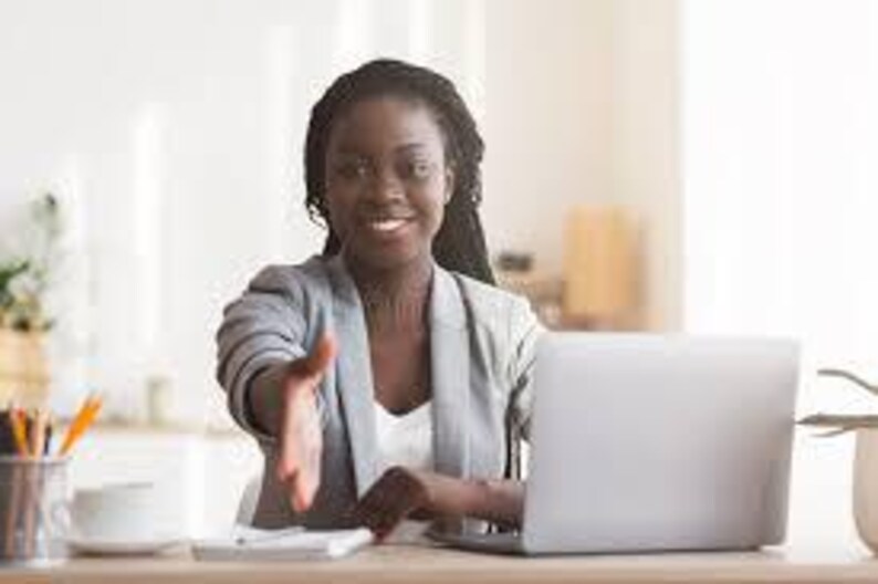 May include: A woman in a gray blazer extends her hand towards the viewer. She is seated at a desk with a laptop, calculator, and a cup of coffee. A pencil holder and a plant are also visible.