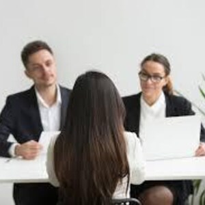 May include: A business meeting with three people seated at a white table. Two people are seated across from a person with long brown hair. A laptop, a cup of coffee, and papers are on the table. The people are wearing business attire.