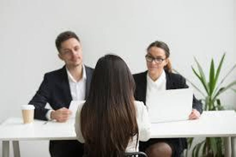 May include: A business meeting with three people seated at a white table. Two people are seated across from a person with long brown hair. A laptop, a cup of coffee, and papers are on the table. The people are wearing business attire.