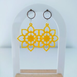 Peut inclure: Une paire de boucles d'oreilles jaunes vif en forme de fleur. Chaque boucle d'oreille présente un motif ajouré détaillé avec des pétales en forme de cœur. Les boucles d'oreilles sont suspendues à de petits cerceaux argentés et sont présentées sur un présentoir blanc avec une base en bois.