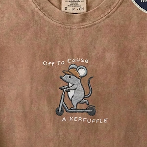 May include: Espresso-coloured t-shirt featuring an embroidered mouse on a scooter, wearing a hat. The text "Off To Cause A Kerfuffle" is embroidered below the mouse. The shirt has a crew neck and a Comfort Colors label.