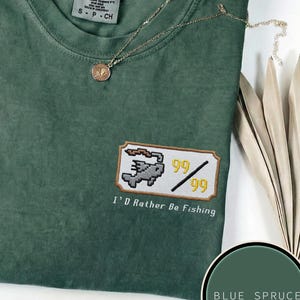 Pode incluir: Camiseta azul abeto com um patch bordado que diz "I'd Rather Be Fishing". O patch apresenta um peixe pixelado e os números "99". Um colar de ouro e um chapéu marrom também são visíveis.