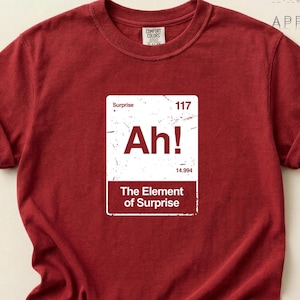 May include: A dark red short-sleeved t-shirt featuring a white rectangular graphic designed like a periodic table element. The graphic displays the text Surprise, 117, Ah!, 14.994, and The Element of Surprise. The shirt has a crew neckline.