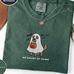 May include: A dark green t-shirt featuring an embroidered graphic of a dog wearing a white ghost costume and holding an orange jack-o'-lantern bucket. The text "WE HAUNT AT DAWN" is embroidered below the dog, surrounded by small black bats.