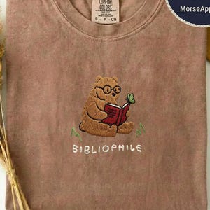 Puede incluir: Una camiseta de cuello redondo color espresso con un bordado de un oso pardo con gafas leyendo un libro rojo con una mariposa verde encima. El texto "BIBLIOPHILE" está bordado en color blanco debajo de la ilustración del oso.