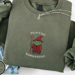 Pode incluir: Uma camisola de gola redonda verde-azeitona com um bordado de um sapo verde vestido com uma túnica e um chapéu de feiticeiro vermelhos, segurando um cajado de madeira. O texto "DELULU BUT DANGEROUS" está bordado a branco à volta.