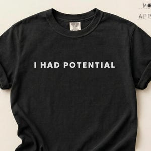 Pode incluir: Uma caneca de cerâmica branca com uma alça preta e o texto "But First, Coffee" em negrito preto. A caneca está sobre uma superfície de madeira. A caneca tem aproximadamente 10 cm de altura.