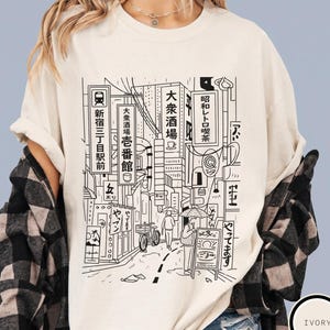 Puede incluir: Camiseta de color marfil con una ilustraci&oacute;n en blanco y negro de una escena callejera japonesa. El dise&ntilde;o incluye texto japon&eacute;s, edificios y personas. La camiseta es de Morse Apparel.