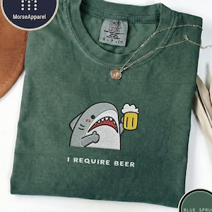 Puede incluir: Una camiseta de cuello redondo de color verde oscuro con un bordado de un tiburón gris sosteniendo una jarra de cerveza amarilla. Debajo del tiburón, el texto I REQUIRE BEER aparece en letras blancas. La camiseta tiene una etiqueta Comfort Colors.