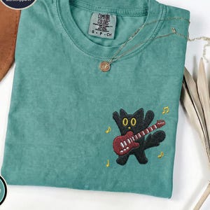 May include: A seafoam green t-shirt featuring an embroidered black cat playing a red electric guitar on the lower right side. The shirt has a crew neckline with a Comfort Colors label and is accented by small yellow musical note embroidery.