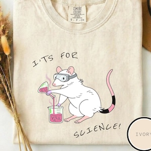 May include: Ivory t-shirt featuring a cartoon rat in goggles, pouring liquid into a beaker. The text "IT'S FOR SCIENCE!" is printed on the shirt. The shirt is a Comfort Colors brand.
