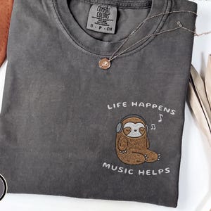 May include: A charcoal grey t-shirt featuring an embroidered graphic of a brown sloth wearing headphones with the text "LIFE HAPPENS MUSIC HELPS" above and below the sloth. The shirt has a crew neckline and a Comfort Colors brand label inside.