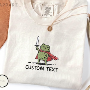 May include: An ivory t-shirt featuring an embroidered green frog wearing a red cape and holding a silver sword. Below the frog, the text reads CUSTOM TEXT. The shirt has a Comfort Colors label at the neck and is paired with a gold pendant necklace.