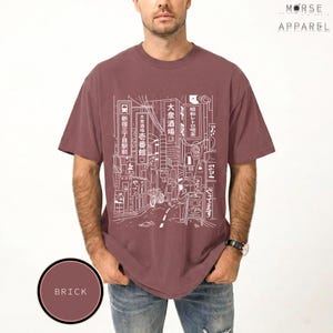 Puede incluir: Camiseta de color ladrillo con un dibujo lineal blanco de una escena callejera japonesa, con letreros y edificios. La camiseta tiene mangas cortas y cuello redondo. La marca "MORSE APPAREL" est&aacute; impresa en la parte superior derecha.
