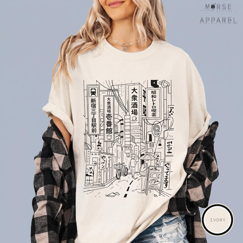 Puede incluir: Camiseta de color marfil con una ilustraci&oacute;n en blanco y negro de una escena callejera japonesa. El dise&ntilde;o incluye texto japon&eacute;s, edificios y personas. La camiseta es de Morse Apparel.