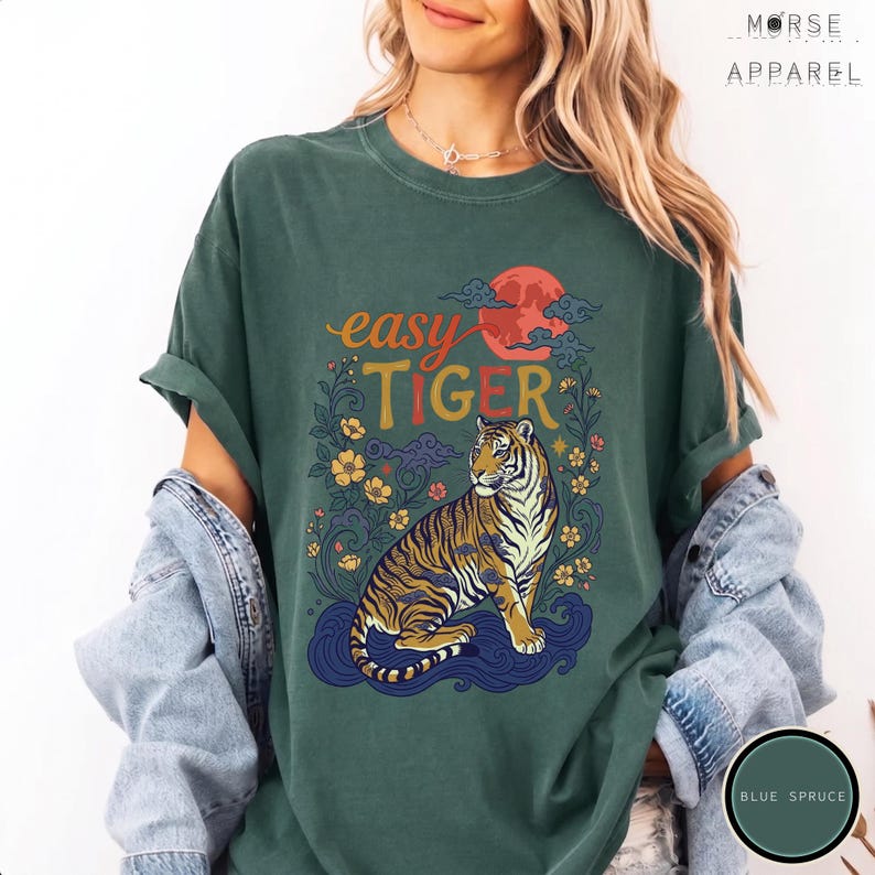 Puede incluir: Camiseta verde oscuro con las palabras "easy TIGER" en naranja y amarillo, con una ilustraci&oacute;n de un tigre. El tigre est&aacute; sentado sobre agua azul con un sol rojo y detalles florales. La camiseta es de Morse Apparel.