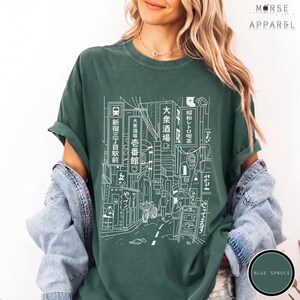 Puede incluir: Una camiseta verde oscuro con un dibujo lineal blanco de una escena callejera japonesa. El dise&ntilde;o incluye edificios, letreros y personas. El texto de la camiseta incluye caracteres japoneses y las palabras "MORSE APPAREL" y "BLUE SPRUCE".