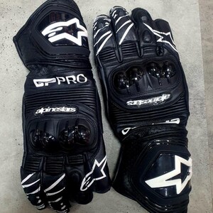 Könnte beinhalten: Paar schwarze Motorradhandschuhe aus Leder mit weißen Akzenten. Die Handschuhe tragen den Schriftzug "GP PRO" und den Markennamen "Alpinestars". Sie haben Knöchelschutz und ein Sternlogo.