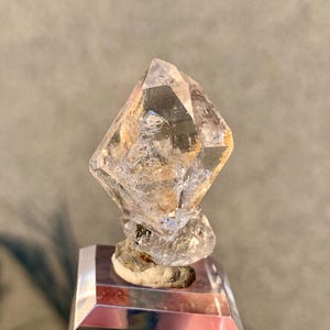 Puede incluir: Un cristal de cuarzo transparente con forma de diamante, montado sobre una base plateada. El cristal presenta inclusiones internas y un aspecto natural sin pulir. La base tiene un diseño geométrico, que refleja la luz. Altura aproximada: 5 cm.