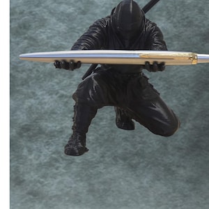 May include: A black ninja figurine in a crouching pose, holding a silver and gold pen. The ninja is wearing a black hooded outfit and has a sword on its back. The pen is held horizontally, appearing to be the focus of the figurine.
