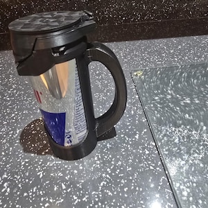 May include: A black 3D-printed mug with a handle, designed to hold a Red Bull can. The mug has a lid and a silver interior, with the Red Bull can visible inside. The mug is sitting on a speckled grey surface.
