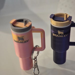 May include: Two miniature Stanley-style travel mugs, one pink and one dark blue. The pink mug has a silver keychain attached. Both mugs have a handle, a lid, and the Stanley logo. The pink mug has a yellow accent, and the blue mug has a gold accent.