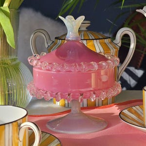 Puede incluir: Un bombonera de cristal rosa con borde con volantes y tapa en forma de flor. El plato se asienta sobre una base de pedestal. Una tetera y una taza de té a rayas amarillas y blancas están en el fondo. El tema general es la decoración vintage.