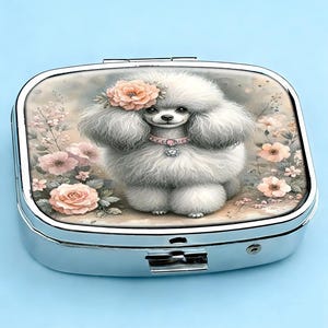 K&ouml;nnte beinhalten: Eine silberne Pillendose mit Klappdeckel, mit einer charmanten Illustration eines flauschigen wei&szlig;en Pudels, der mit einem Blumenkranz und einem rosa Halsband geschm&uuml;ckt ist. Der Hintergrund umfasst zarte rosa Blumen und Bl&auml;tter.
