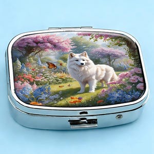 K&ouml;nnte beinhalten: Eine silberne Pillendose mit Klappdeckel. Die Oberseite zeigt eine farbenfrohe Illustration eines wei&szlig;en Hundes in einem Garten mit Blumen und Schmetterlingen. Die Dose hat eine rechteckige Form mit abgerundeten Ecken und einem sicheren Verschluss.