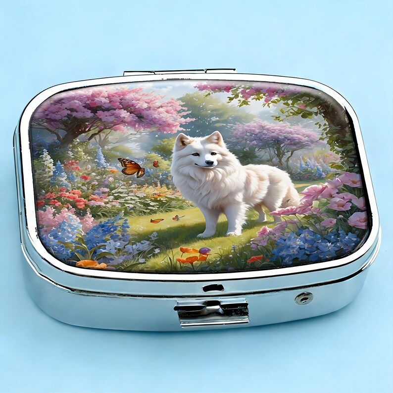 K&ouml;nnte beinhalten: Eine silberne Pillendose mit Klappdeckel. Die Oberseite zeigt eine farbenfrohe Illustration eines wei&szlig;en Hundes in einem Garten mit Blumen und Schmetterlingen. Die Dose hat eine rechteckige Form mit abgerundeten Ecken und einem sicheren Verschluss.