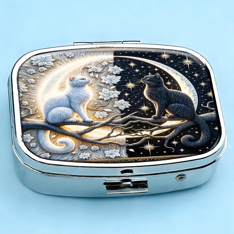 K&ouml;nnte beinhalten: Silberne Pillendose mit detailliertem Design, das eine wei&szlig;e Katze auf einem Ast vor einem leuchtenden Mond und eine schwarze Katze vor einem Sternenhimmel zeigt. Die Dose hat einen Klappdeckel und einen sicheren Verschluss.