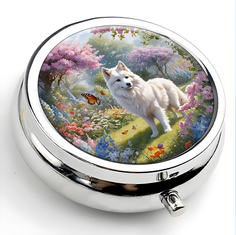 K&ouml;nnte beinhalten: Eine runde, silberne Pillendose mit einem detaillierten, farbenfrohen Bild auf dem Deckel. Das Bild zeigt einen wei&szlig;en Hund in einem lebendigen Garten mit Schmetterlingen, Blumen und B&auml;umen. Die Pillendose hat eine gl&auml;nzende, metallische Oberfl&auml;che und einen kleinen Verschluss.