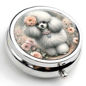 K&ouml;nnte beinhalten: Eine silberne Pillendose mit einer detaillierten Illustration eines flauschigen wei&szlig;en Pudels mit rosa Halsband und Blumenkrone. Der Pudel ist von rosa Blumen umgeben. Die Pillendose hat einen Klappdeckel und einen kleinen Verschluss.