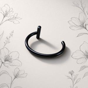 May include: A black, circular body jewellery piece featuring a smooth, curved band that forms an open hoop shape. One end of the band terminates in a small, cylindrical post that stands perpendicular to the main circular structure of the jewellery.