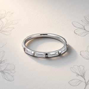 Può includere: Un anello a fascia in metallo color argento con una serie di pietre trasparenti a taglio baguette rettangolare incastonate orizzontalmente lungo la circonferenza. L'anello presenta una finitura lucida e riflettente con un profilo sottile e uniforme.