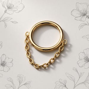 Könnte beinhalten: Ein goldfarbener, kreisförmiger Ring mit einer zarten, befestigten Kette, die von den Seiten des Reifens herabhängt. Das Schmuckstück hat eine polierte, reflektierende Metalloberfläche und ein nahtloses, durchgehendes Design mit Klappverschluss.