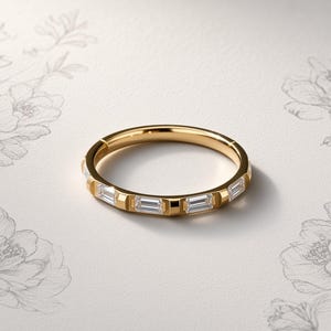 Könnte beinhalten: Ein goldfarbener Ring mit einer Reihe klarer, rechteckiger Baguette-Steine, die bündig in das Band eingelassen sind. Das Metall hat eine polierte, reflektierende Oberfläche und das Design zeigt einen sichtbaren Scharniermechanismus.