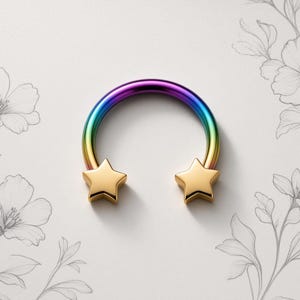 Pode incluir: Uma joia corporal circular em forma de ferradura com um gradiente de arco-íris vibrante ao longo da barra curva. As duas extremidades do anel são rematadas com estrelas de cinco pontas em tom dourado polido e simétricas.