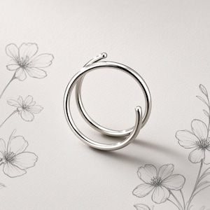 Könnte beinhalten: Ein polierter Ring in Silberoptik mit einem Bypass-Design, bei dem sich zwei Bänder überlappen, ohne sich zu berühren. Das Metall hat eine glatte, reflektierende Oberfläche und bildet eine schlichte, kreisförmige Form.