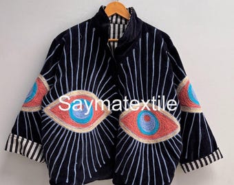 May include: A black jacket featuring large embroidered eye motifs on the front and sleeves, with light blue and orange details. The garment has a high collar, three-quarter length sleeves with black and white striped cuffs, and the text Saymatextile.