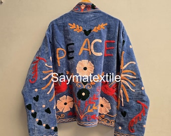 May include: A blue jacket featuring colorful embroidery with the word PEACE across the back. The design includes floral patterns, bird motifs, and heart shapes in orange, red, pink, and black thread. The text Saymatextile is visible on the jacket.