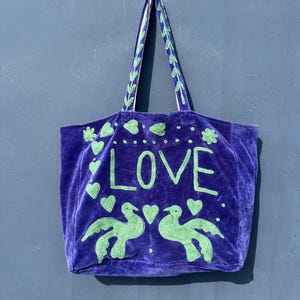 May include: A deep purple tote bag with the word "LOVE" in large, light green letters. The bag features green heart and bird designs. The handles are purple with green embroidered accents. The bag is made of a soft, velvety material.