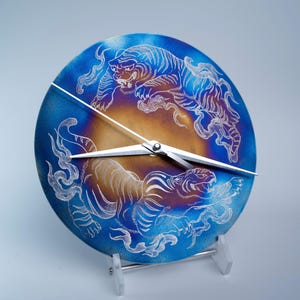 Puede incluir: Un reloj redondo con una esfera degradada azul y dorada con diseños de tigres grabados. El reloj tiene manecillas blancas y está montado sobre un soporte transparente. Los tigres están detallados con líneas blancas.