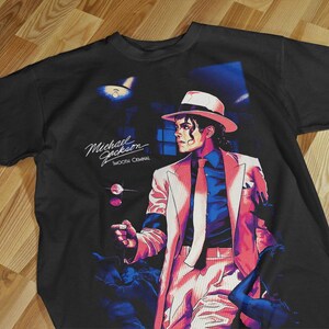 Puede incluir: Camiseta negra con un gráfico de Michael Jackson con traje blanco y sombrero fedora. La imagen tiene un estilo retro con vibrantes tonos rosa, azul y rojo. El texto "Michael Jackson Smooth Criminal" está impreso en la camiseta.