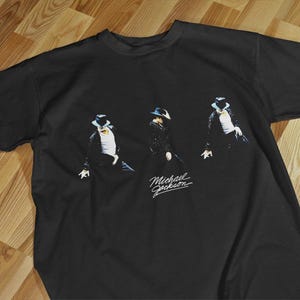 Puede incluir: Camiseta negra con un gráfico de Michael Jackson en una pose de baile. El diseño muestra tres figuras con trajes y sombreros negros, con el nombre del artista en escritura cursiva. La camiseta se exhibe sobre una superficie de madera.