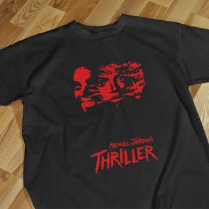 Puede incluir: Camiseta negra con un gráfico rojo de caras y el texto "Michael Jackson's Thriller". El diseño está centrado en la camiseta. La camiseta está hecha de un material suave.