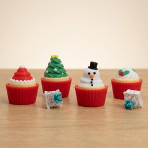 May include: Four Christmas-themed cupcake decorations. One is a Santa hat, another a Christmas tree, a snowman, and a cupcake with holly. Each is in a red cupcake case. Two small, clear, square objects are in front of the cupcakes.
