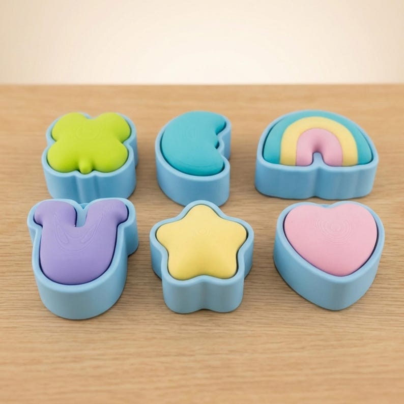 May include: Six colourful, shaped soap bars. The shapes include a four-leaf clover, crescent moon, rainbow, horseshoe, star, and heart. Each soap is a different colour and sits in a light blue container. The background is a light brown wooden surface.