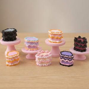 May include: A collection of miniature cakes in assorted colours, including pink, black, purple, and orange, displayed on small pink cake stands. The cakes are decorated with intricate frosting details, creating a whimsical and charming appearance. The cakes are arranged on a light brown surface.
