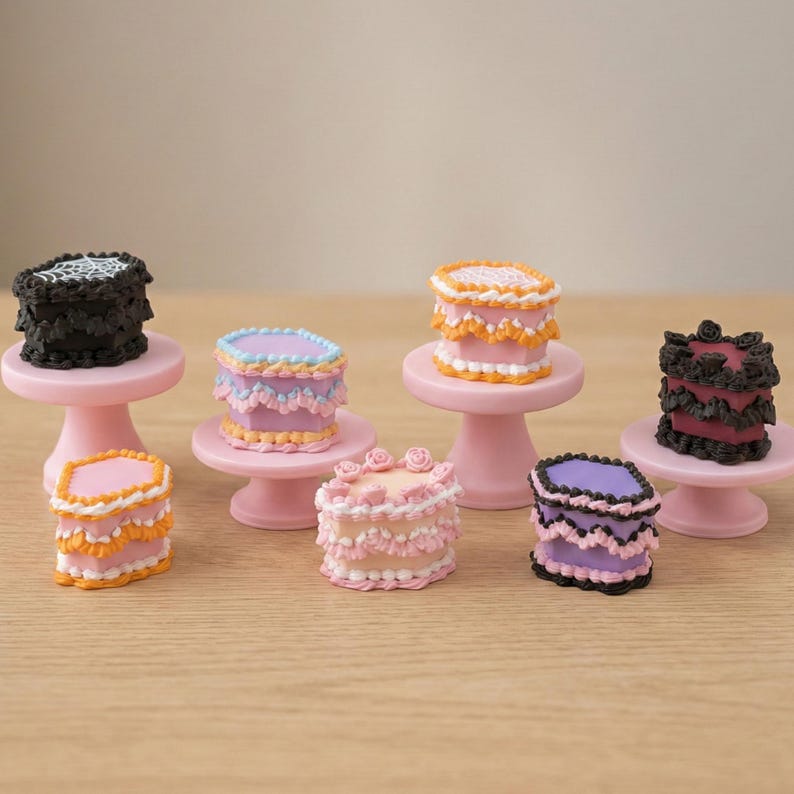 May include: A collection of miniature cakes in assorted colours, including pink, black, purple, and orange, displayed on small pink cake stands. The cakes are decorated with intricate frosting details, creating a whimsical and charming appearance. The cakes are arranged on a light brown surface.